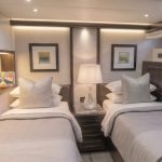 Httpswww Andyquinones Net luxurious yacht cabin with two single beds soft gray bedding and matching pillows facing each other across a nightstand A mirror lamps framed art and decorative objects line the wall behind the beds porthole windows dot the opposite wall